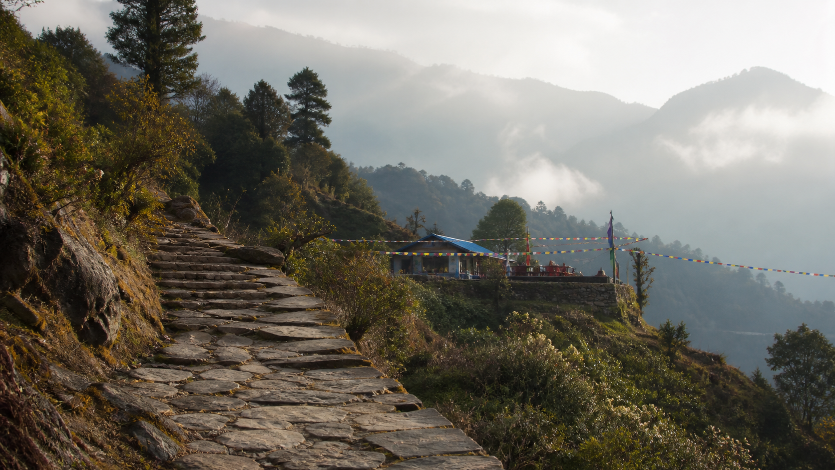 Himalaya-Vorland in Nepal mit Steinweg, Gebetsfahnen, Terrassenhängen und fernen Schneebergen mit ruhiger Stimmung im Beitrag.